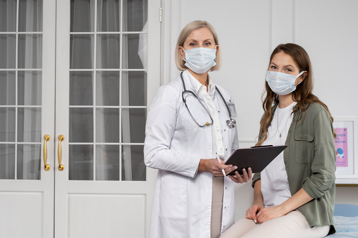 Muayene odasında tıbbi maske takmış, elinde dosya tutan doktor ve yanındaki maskeli kadın hasta.
