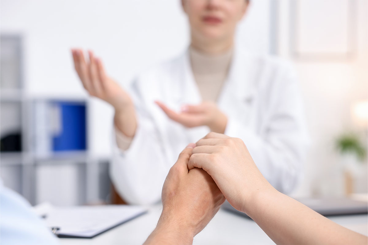 Masada ellerini üst üste koyarak birbirine destek olan çift ve karşılarında konuşan kadın doktor.